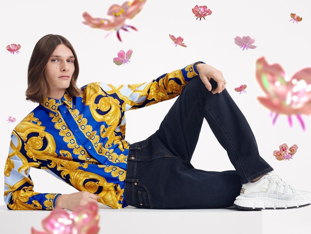Campaign image: a man in a blue and gold baroque-print shirt and dark jeans reclines on a white surface, surrounded by floating pink flowers.