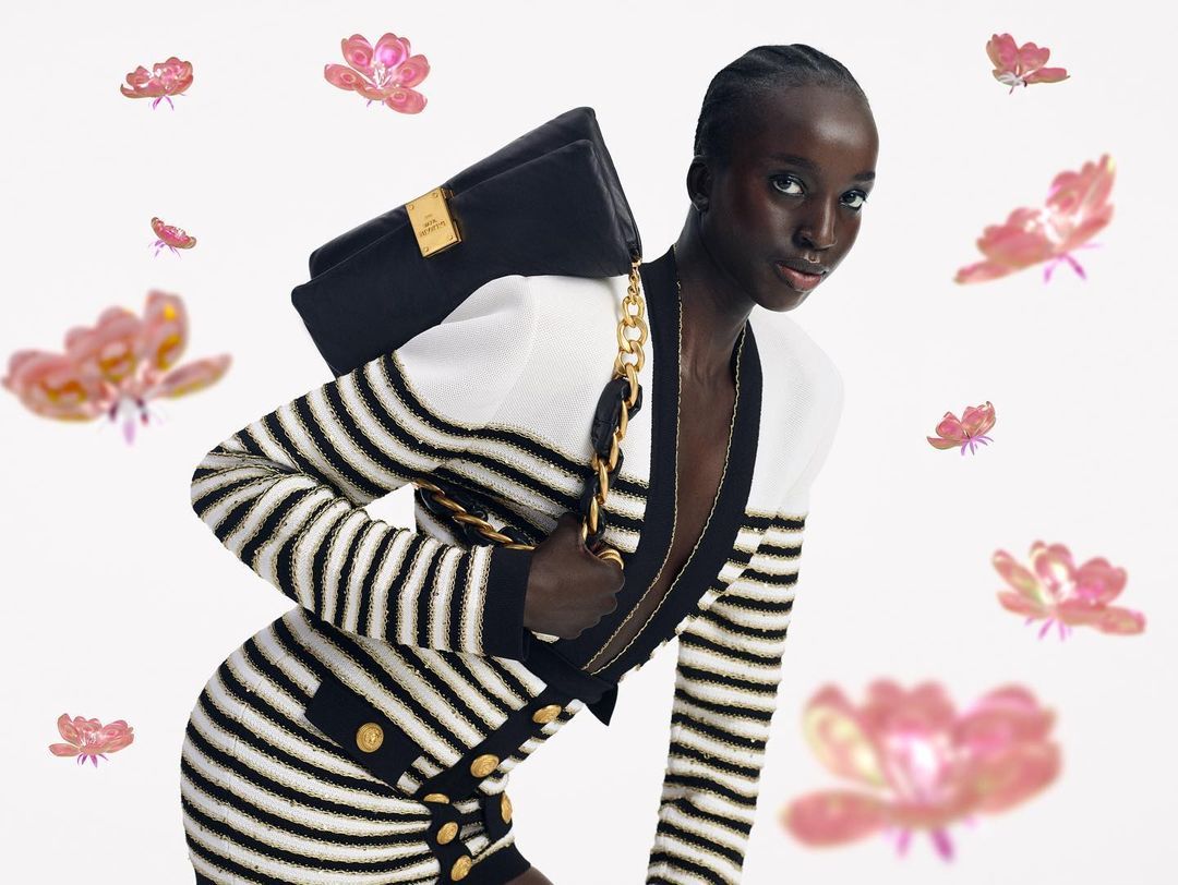 Campaign image: a woman in a black and white striped knit jacket carries a black bag on a gold chain, against a white background with floating pink flowers.