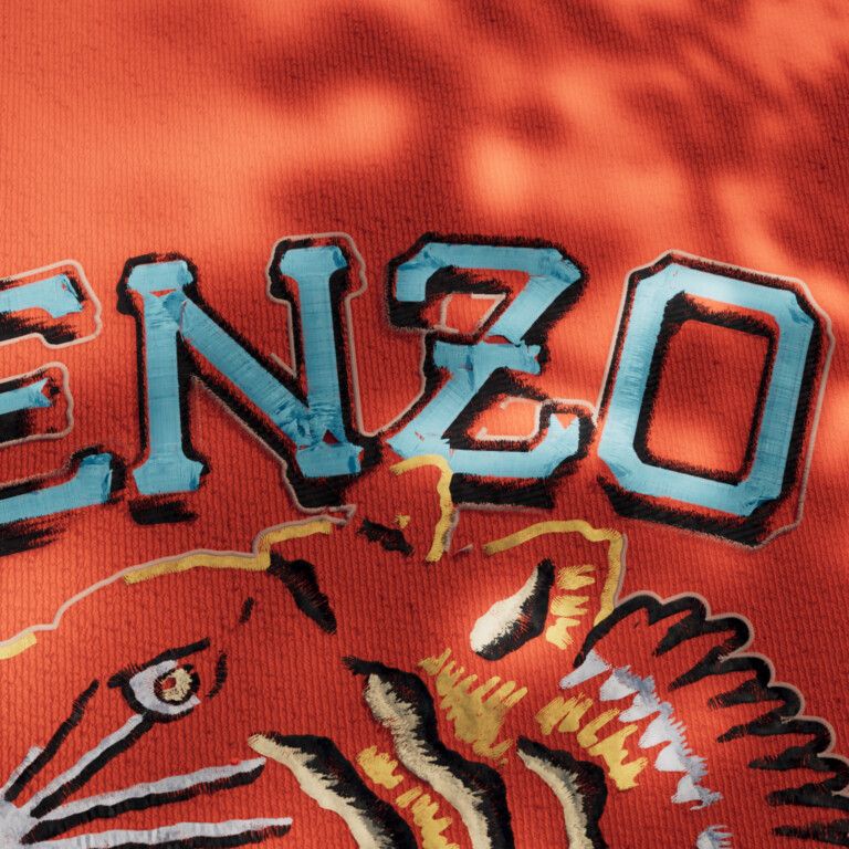 Orange knitted fabric in dappled shadow, with the word KENZO in raised light-blue stitched letters above part of a stitched tiger head in black, white and yellow.