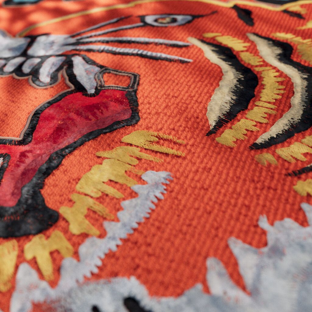 Close-up of a stitched tiger face on orange knit: an eye, white whiskers, a red tongue outlined in black, and black, cream and yellow stripes.