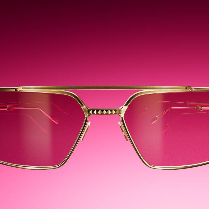 Front view of gold metal sunglasses with pink lenses and small studs on the bridge, against a pink to dark red gradient.