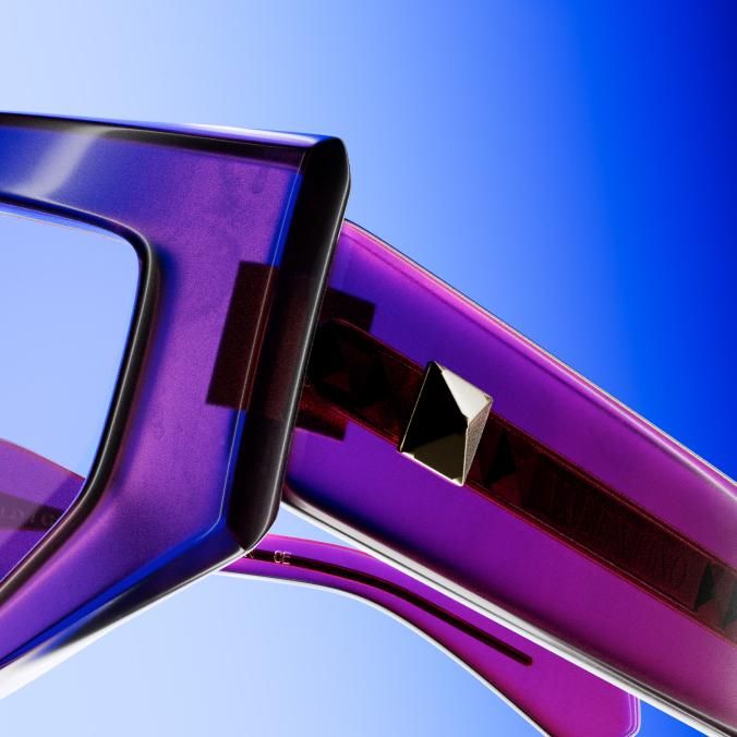 Close-up of a translucent violet frame and temple with a gold stud, against a blue gradient.