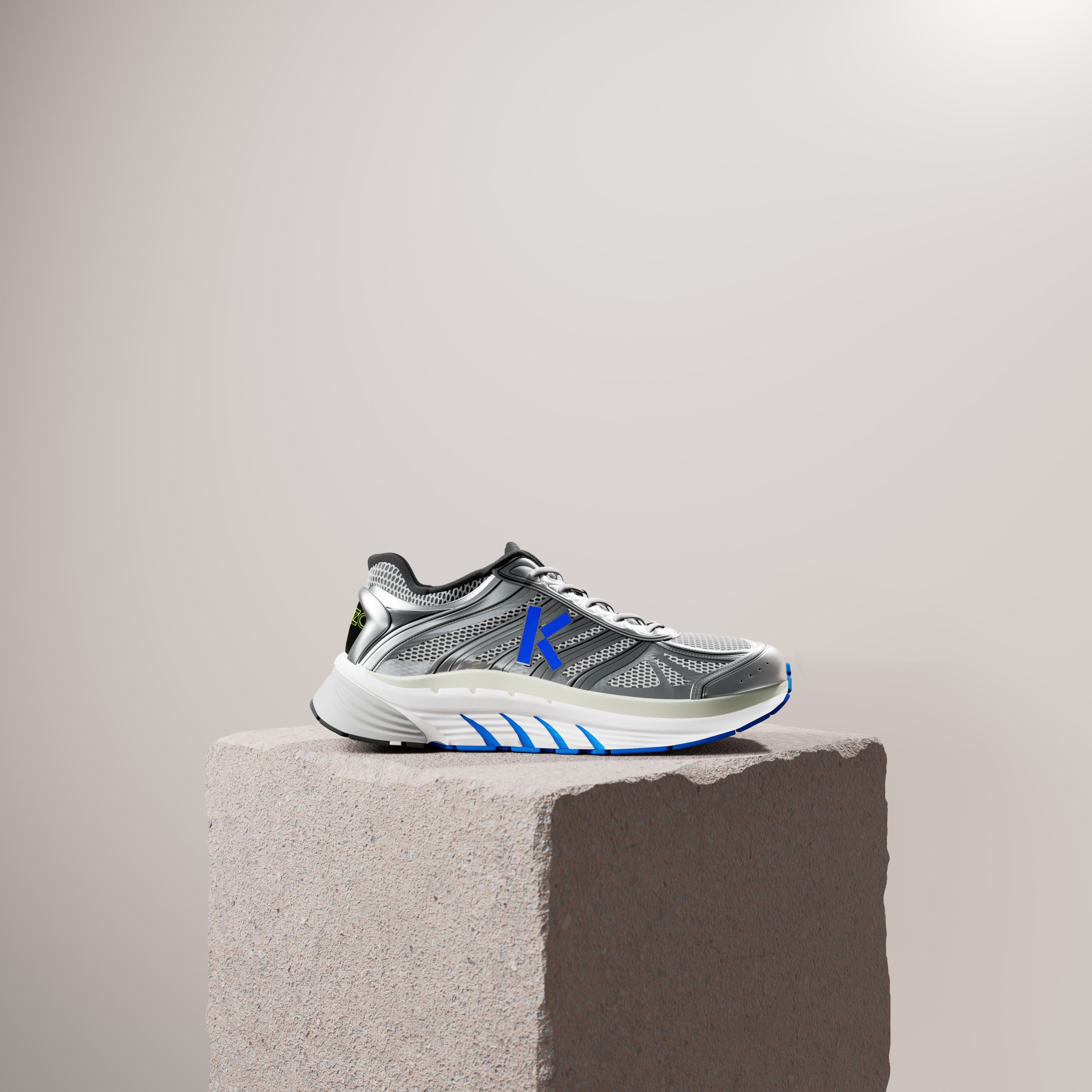 A silver running shoe with a blue K and blue sole details on a rough stone plinth against a pale beige wall.