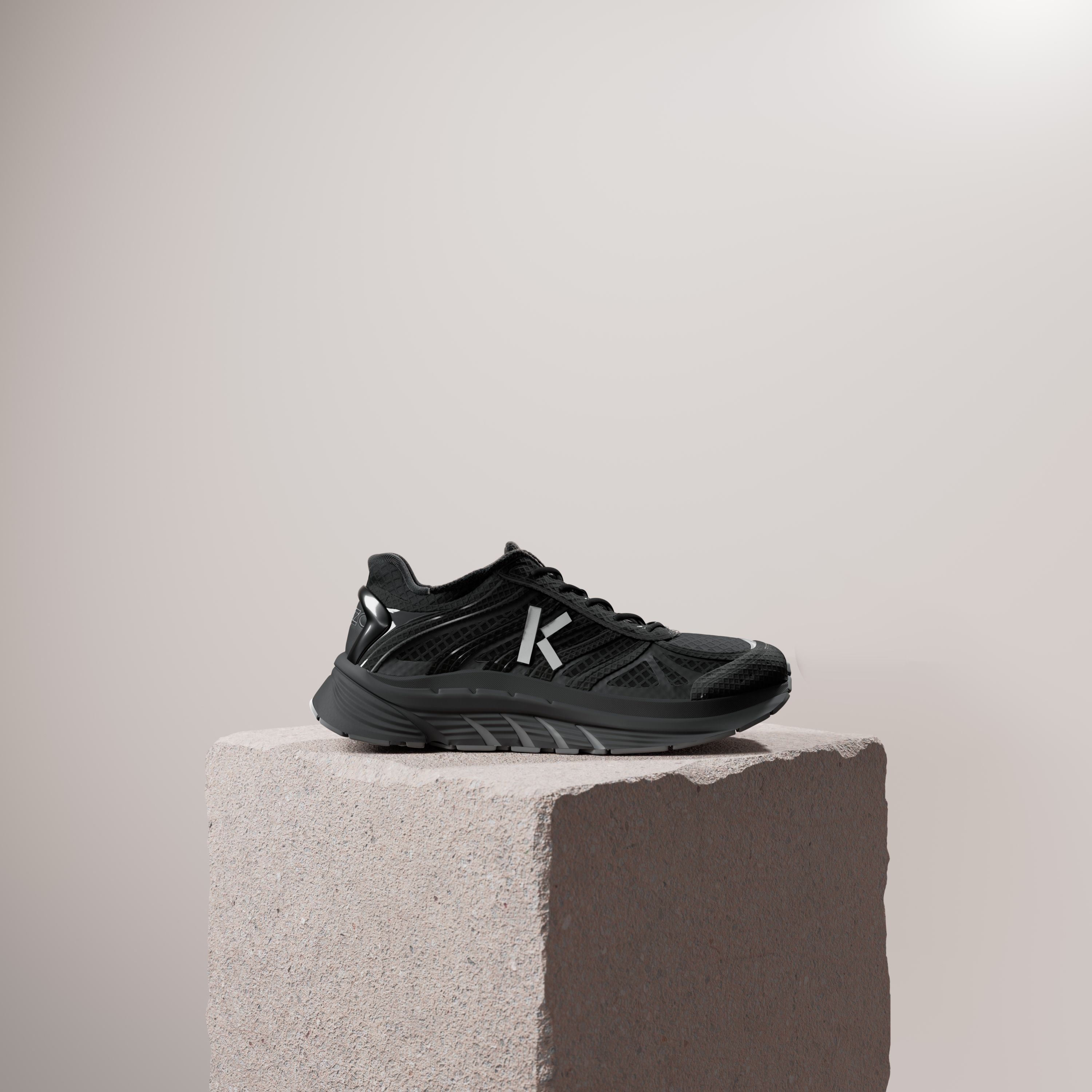 An all-black running shoe with a white K on a rough stone plinth against a pale beige wall.